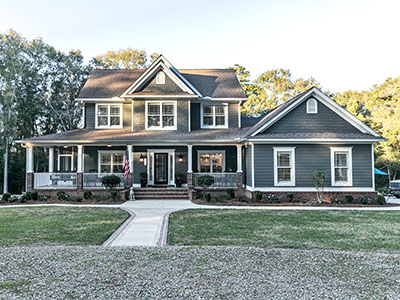 Front view of a large two story blue gray house with wood and vinyl siding with an expansive porch, sidewalk and manicured lawn.